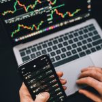 Person analyzing stock market data with a laptop and smartphone showing trading graphs.